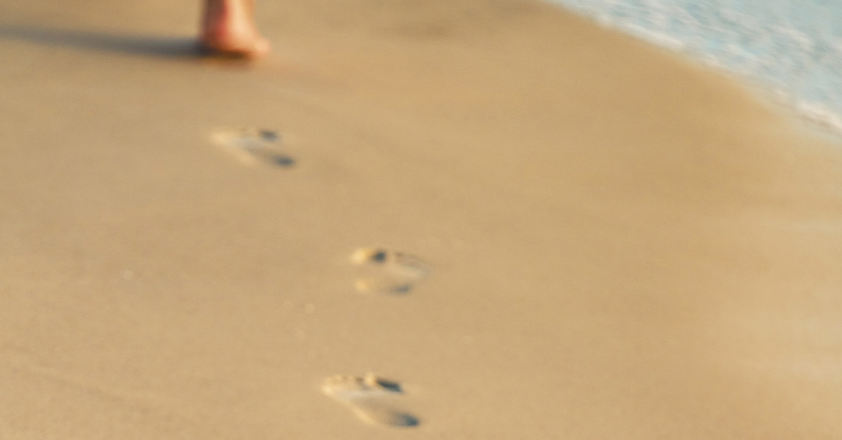 Footprints in sand near shoreline with person walking along beach.