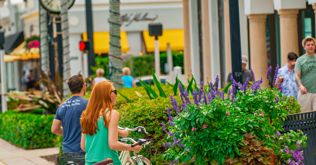 People walking and biking along landscaped downtown street with flowers and shops.