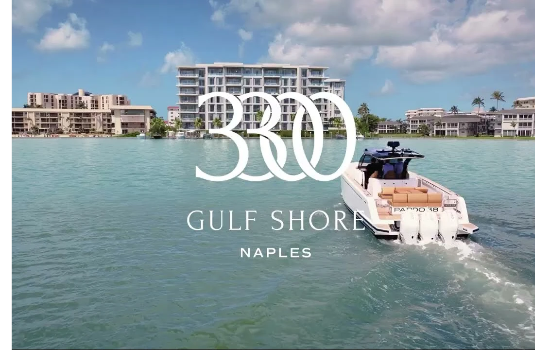 Waterfront condo building with marina, boats, and palm trees under blue sky.