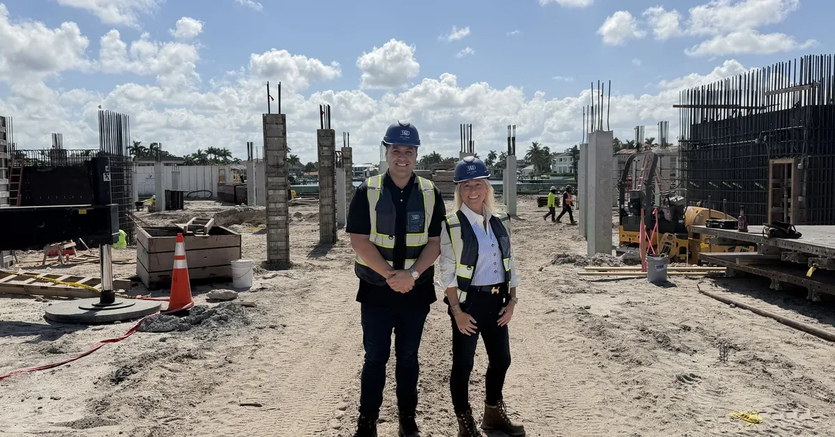 Two real estate professionals in hard hats and safety vests tour an active waterfront construction site with concrete columns and rebar framework.
